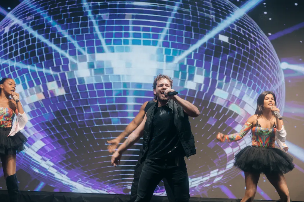 Cantor de preto diante do globo espelhado em LED, artistas de tutu dançam