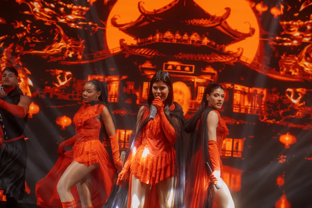 Três artistas de vermelho cantam e dançam diante do cenário oriental em LED