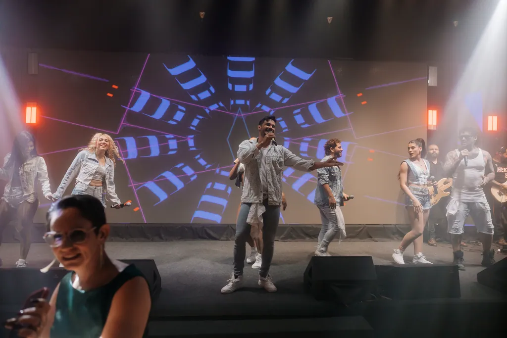 Artistas de branco e jeans no palco azul; convidada canta junto na frente